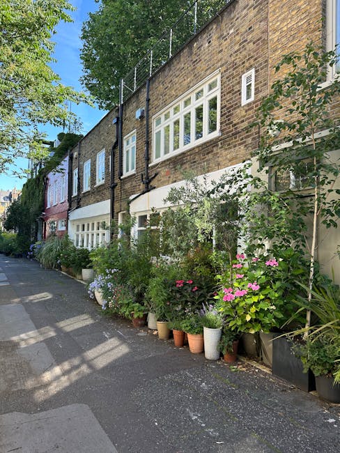 A row of potted plants and flowers, including pink hydrangeas, purple petunias, and green foliage, arranged along the base of a brick residential building with white-framed windows. The plants are in 