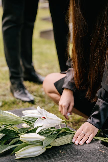 A person dressed in dark clothing, with long brown hair, is kneeling on the ground, placing white lilies with long green stems and leaves onto a grave or memorial stone. The lilies appear fresh and we