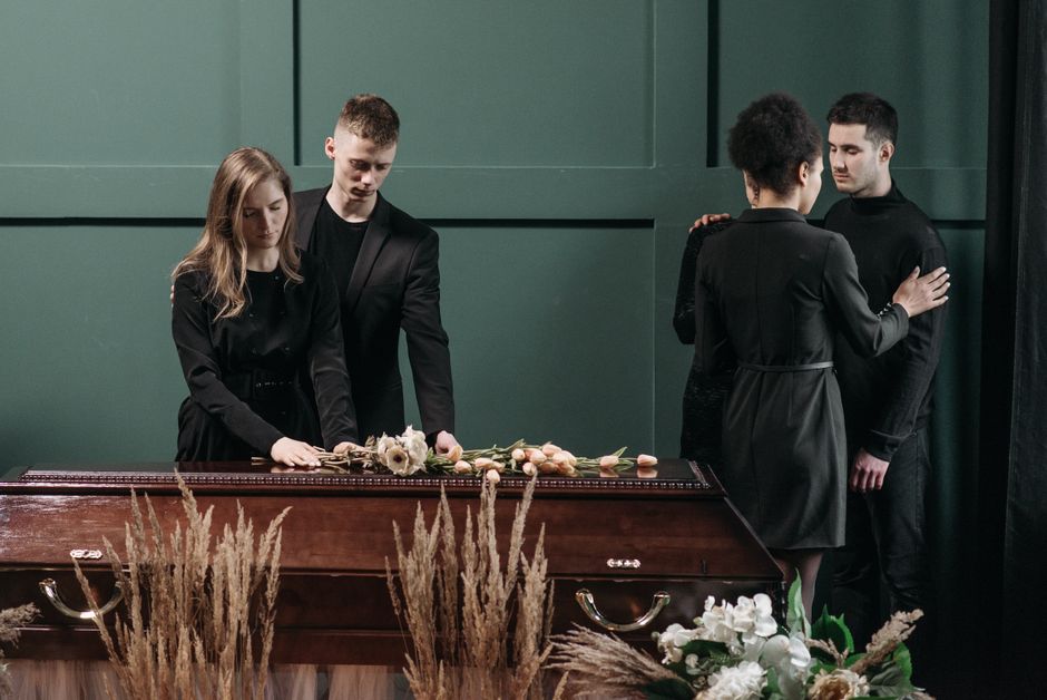 Two women and two men dressed in black mourning attire standing near a casket during a funeral service. The woman on the left is resting her hands on the closed, polished wooden casket which is decora